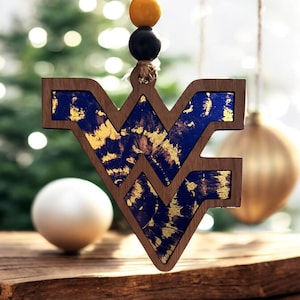 May include: Wooden Christmas ornament in the shape of the letters WV, with a dark wood frame and blue and gold interior. The ornament hangs from a string with yellow and black beads. A white sphere and gold ornament are in the background.
