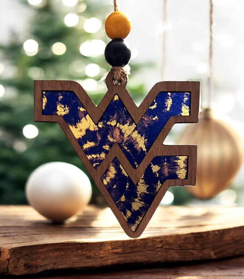 May include: Wooden Christmas ornament in the shape of the letters WV, with a dark wood frame and blue and gold interior. The ornament hangs from a string with yellow and black beads. A white sphere and gold ornament are in the background.