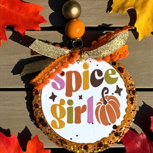 May include: A circular ornament with the words "Spice Girl" in various colours and a pumpkin illustration. The ornament has a glittery edge, gold ribbon, and orange pom-poms. It hangs from a gold cord and beads. Autumnal decoration.