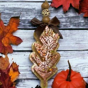 May include: A leaf-shaped ornament with a copper glitter design and gold outline, ideal for autumn decor. It features a brown bow, leopard print and brown beads, and a hanging string. The background includes red and orange autumn leaves and a small pumpkin.
