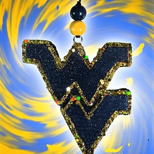 May include: A navy blue glittered pendant in the shape of the West Virginia University logo, outlined in gold glitter. The pendant hangs from a black cord with navy and yellow beads. The background is a swirl of blue and yellow.