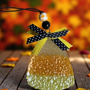 May include: A candy corn-shaped air freshener with a black and white polka dot bow and yellow trim. The air freshener has a string for hanging and a decorative bead. The background is a blurred autumn scene.