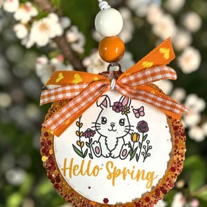 Puede incluir: Adorno redondo y decorativo con temática primaveral. El adorno presenta una ilustración de un conejito blanco rodeado de flores y el texto "Hello Spring". Tiene un borde de purpurina naranja, un lazo a cuadros naranja y blanco y una cuerda de cuentas blancas y naranjas.