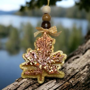 May include: A decorative maple leaf ornament with a gold glitter outline and a brown and copper glitter interior. The ornament is suspended by a gold string and adorned with wooden beads and a gingham ribbon. The background is a blurred view of a lake and trees.