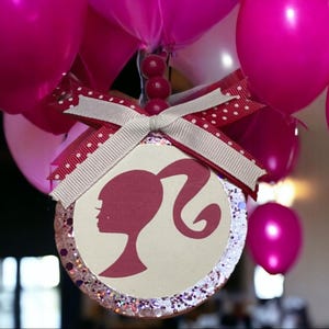 May include: A pink and white round bauble with a glitter finish and a silhouette of a Barbie doll. The bauble is decorated with a white ribbon with pink polka dots.