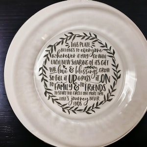 May include: A white ceramic plate with a black inscription that reads "This plate belongs to everyone wherever it may go with each new sharing of its gift the love & blessings grow so fill it up & pass it on to family & to friends to start the circle one more time love's journey never ends".