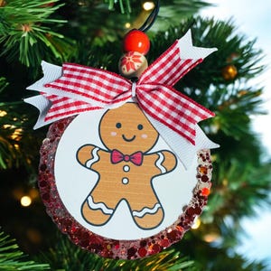 May include: A round Christmas ornament featuring a gingerbread man illustration with a red bow tie. The ornament has a red and white checkered ribbon and is surrounded by red glitter. It hangs from a black string.