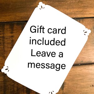May include: White gift card with decorative corner cutouts. The card includes the text "Gift card included Leave a message" in black font. The card is set against a wooden background.