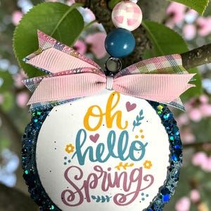 Puede incluir: Adorno redondo blanco con las palabras "oh hello Spring" en escritura colorida. El adorno tiene un borde azul brillante y está adornado con un lazo rosa y cuentas decorativas. Colgando de una rama de árbol.