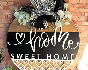 May include: A round wooden door hanger with a black and white chevron pattern at the bottom. The top half of the door hanger is painted black with the word "home" written in white cursive script. The words "sweet home" are written in white block letters below the cursive word "home". A black and white striped bow with a burlap bow is attached to the top of the door hanger.