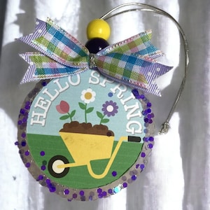 May include: A purple and white polka dot bauble with a yellow and purple bow. The bauble has a blue background with the text "Hello Spring" and a cartoon image of a wheelbarrow with flowers.