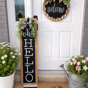 May include: A welcoming front porch scene with a wooden sign saying "oh, HELLO", a round "welcome" sign, and a "Hello" doormat. The scene includes potted flowers and a watering can, creating a warm, inviting atmosphere for visitors.
