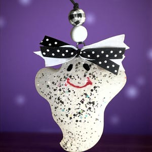 Könnte beinhalten: Glitzernder, weißer Geist-Ornament mit einer schwarz-weiß gepunkteten Schleife und einem roten, lächelnden Mund. Die Halloween-Dekoration hängt an einer schwarzen Schnur und hat eine karierte Perle.