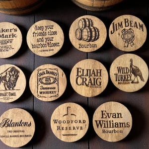 May include: A collection of wooden coasters with various bourbon and whiskey brand logos and slogans, including "Maker's Mark," "Jim Beam," and "Buffalo Trace." The coasters are round and made of light-colored wood.
