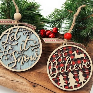 May include: Two wooden Christmas ornaments. One has "Let it Snow" on a blue background with snowflakes. The other has "Believe" on a red and black plaid background with a reindeer and trees. Both have twine hangers.
