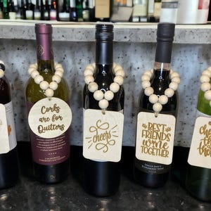 May include: Five wine bottles with wooden bead necklaces and decorative tags. The tags have phrases like "Congratulations," "Cheers," and "Best Friends Wine Together." The bottles are various colors, including red and green.
