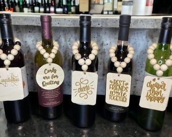 Op de afbeelding: Vijf wijnflessen met houten kralenkettingen en decoratieve labels. De labels bevatten zinnen als "Gefeliciteerd", "Proost" en "Beste vrienden wijn samen". De flessen zijn in verschillende kleuren.
