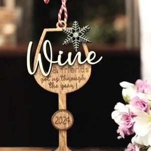 May include: Wooden wine glass ornament with the words "Wine & Friends got us through the year!" and the year 2024. The ornament has a snowflake and is hanging from a red and white string.