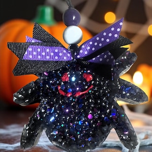 May include: A black glitter spider ornament with a smiling red face, purple and white polka dot bow, and black ribbon. The Halloween decoration has a string for hanging and is set against a blurred background with a pumpkin and spider web.