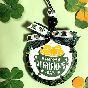 May include: A St. Patrick's Day themed ornament with a green and white bow, a black string, and a white bead with a shamrock design. The ornament features a pot of gold and the text "Happy St. Patrick's Day". Green glitter surrounds the ornament.
