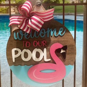 Puede incluir: Un colgador de puerta redondo de madera con el texto "Bienvenido a nuestra piscina" en letras azules y blancas. Un flamenco rosa con gafas de sol flota en un flotador rosa inflable delante de un diseño de ola azul y blanco.
