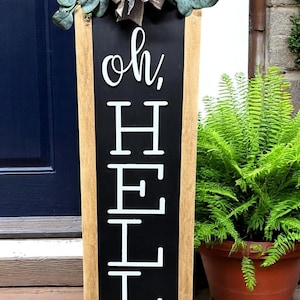 May include: A wooden welcome sign with the words "oh, HELLO" in white lettering on a black background. The sign is framed in light brown wood and adorned with greenery and a decorative bow. A fern in a terracotta pot is visible.