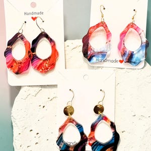 May include: Three pairs of handmade earrings with a colorful, abstract design. The earrings have a teardrop shape with a wavy edge and a gold-colored hook. The earrings are displayed on white cards with the word "Handmade" and a heart symbol.