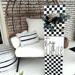 May include: A black and white chequered welcome sign with the words "stay a while" in gold script. The sign is decorated with a bow, greenery, and wooden beads. The sign is displayed on a porch with a wicker chair and white cushions.