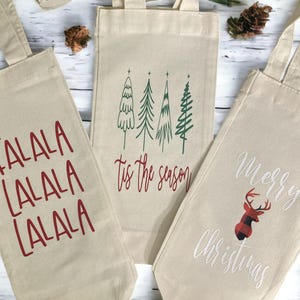 May include: Three beige canvas wine bags with holiday-themed designs. One bag has the text "FALALA" in red, another has green tree illustrations and the text "tis the season", and the third has "Merry Christmas" with a plaid reindeer.