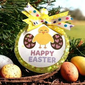 May include: A round Easter decoration featuring a yellow chick and the words "HAPPY EASTER". The ornament has a yellow bow with polka dots. Decorative eggs are in a nest in the foreground.