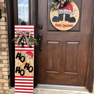 Puede incluir: Un letrero de puerta navideño de madera y un letrero vertical. El letrero redondo de la puerta presenta las piernas de Papá Noel y las palabras "Merry Christmas". El letrero vertical tiene rayas rojas y blancas, las palabras "Ho Ho Ho" y un gorro de Papá Noel.