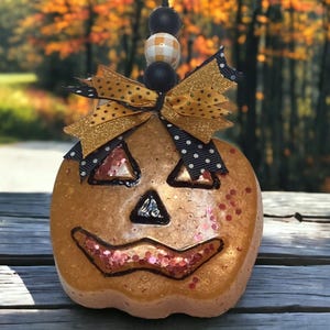 Könnte beinhalten: Dekorative Kürbis-Ornament mit geschnitztem Gesicht, Glitzer und einer Schleife. Der Kürbis ist orange mit schwarzen Umrissen und einer schwarzen Dreiecksnase. Die Schleife ist goldfarben und mit schwarzen Punkten versehen. Das Ornament hängt an einer Schnur.