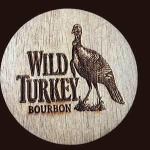 May include: A round wooden coaster with the words "WILD TURKEY BOURBON" in a bold, vintage font. A detailed engraving of a turkey is on the right side. The coaster has a rustic, weathered appearance.
