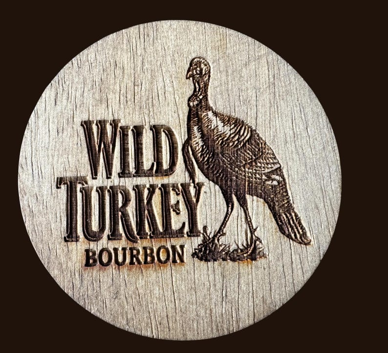 May include: A round wooden coaster with the words "WILD TURKEY BOURBON" in a bold, vintage font. A detailed engraving of a turkey is on the right side. The coaster has a rustic, weathered appearance.