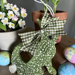 May include: A green, textured bunny ornament with a gingham bow, a white tail, and a pink nose. The bunny is hanging in front of potted flowers and decorated Easter eggs. The ornament is on a wooden surface.