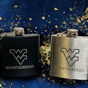 May include: Two stainless steel hip flasks with a silver and black finish. Both flasks feature a  'W' logo and the text 'Mountaineers'.