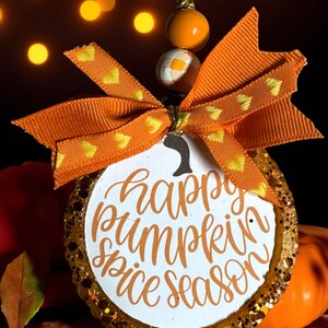 May include: A circular pumpkin-shaped decoration with the words "happy pumpkin spice season" in orange script. It has a gold glitter border and an orange bow with yellow heart details. An orange beaded string is attached for hanging.