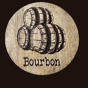 May include: A round wooden coaster with a laser-etched design of bourbon barrels. The word "Bourbon" is etched below the barrels. The coaster is made of light-colored wood and has a dark brown background.