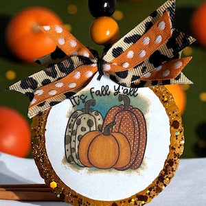 May include: A seasonal ornament with a pumpkin illustration and the text "It's Fall Y'all". The ornament has a gold glitter border and a bow made of leopard and polka dot ribbons. A black and orange bead string is attached for hanging.