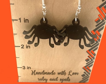 May include: A pair of black spider-shaped earrings with silver hooks. The earrings are approximately 2 inches in length. The background is a light brown surface with the text "Handmade with Love ruby and opals".