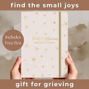 May include: A cream-colored gratitude journal with gold star patterns and the text "DAILY Glimmers GRATITUDE JOURNAL." The journal is held by hands and includes a free pen. The image also includes the text "find the small joys" and "gift for grieving."