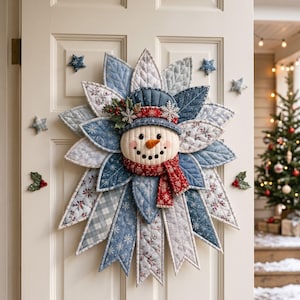 Peut inclure: Une décoration murale en tissu matelassé représentant un bonhomme de neige avec un nez en carotte orange, portant un chapeau bleu et une écharpe rouge à motifs. Le bonhomme de neige est entouré de feuilles en tissu superposées dans des tons de bleu, blanc et gris avec des imprimés hivernaux, agrémentées de petites étoiles et de branches de houx.