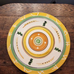 May include: A decorative ceramic plate with a vibrant design. The plate features concentric circles in yellow, orange, and green, with black and white geometric patterns. The outer rim is yellow with white symbols.