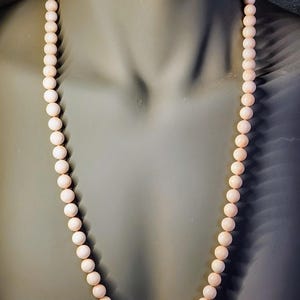 May include: A long necklace made of pale pink coral beads. The beads are round and uniform in size. The necklace is displayed on a mannequin.