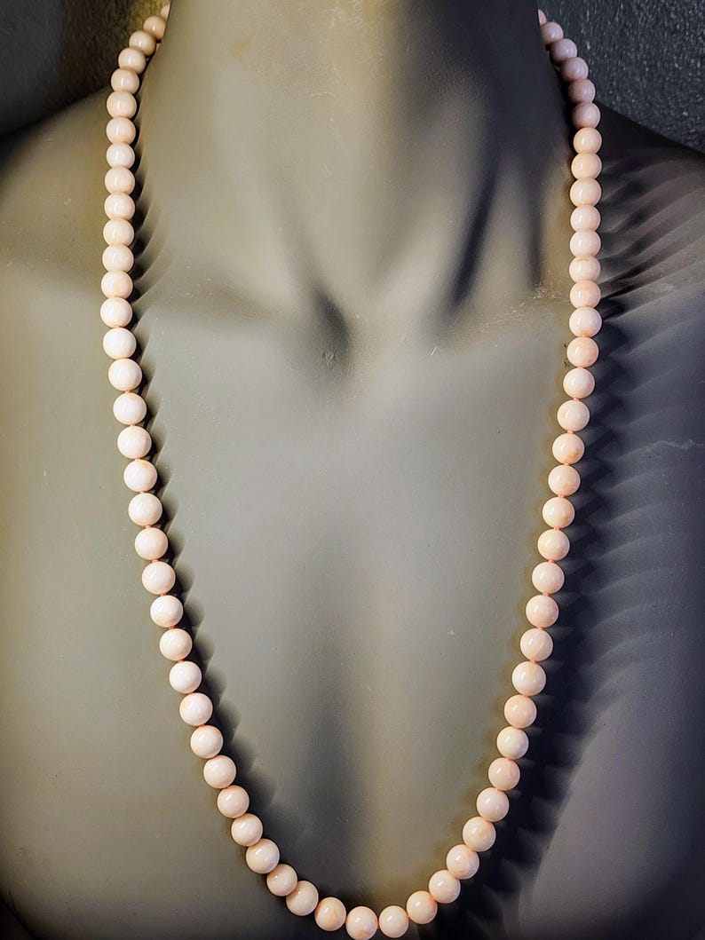 May include: A long necklace made of pale pink coral beads. The beads are round and uniform in size. The necklace is displayed on a mannequin.