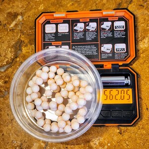May include: A white digital scale with a black display showing a weight of 56.205 grams. A clear plastic container with a string of white beads is on the scale. The scale has a black and orange casing with instructions for using the scale.