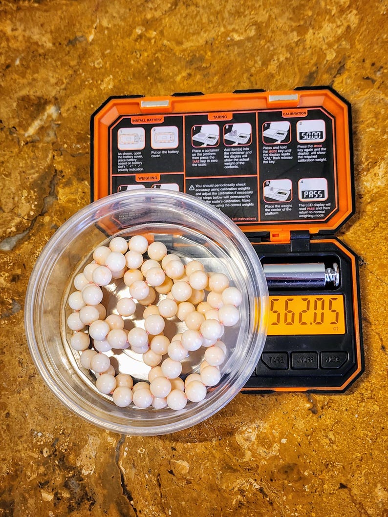 May include: A white digital scale with a black display showing a weight of 56.205 grams. A clear plastic container with a string of white beads is on the scale. The scale has a black and orange casing with instructions for using the scale.