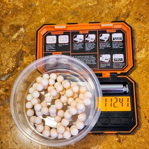 May include: A white digital scale with a black display showing 112.41 grams. The scale is in a black and orange case with instructions for using the scale. A small bowl with a strand of white and pink beads is next to the scale.