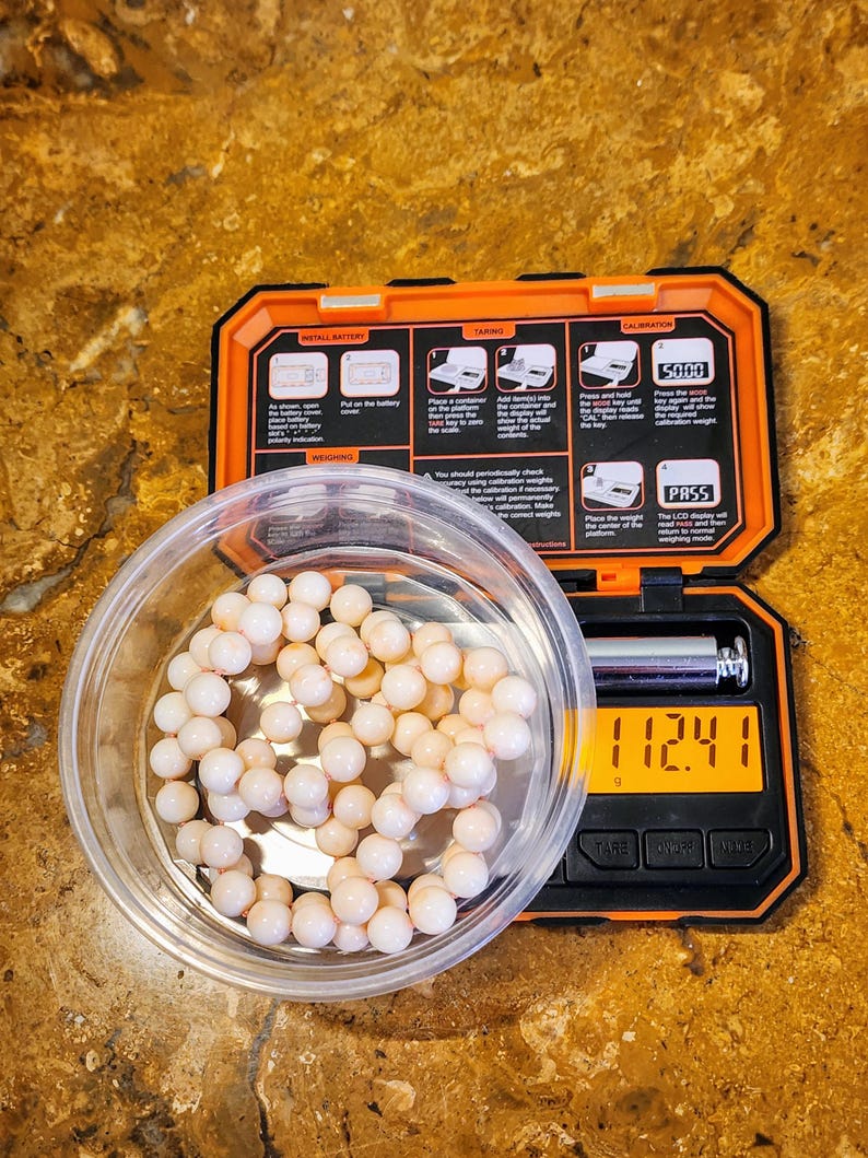 May include: A white digital scale with a black display showing 112.41 grams. The scale is in a black and orange case with instructions for using the scale. A small bowl with a strand of white and pink beads is next to the scale.