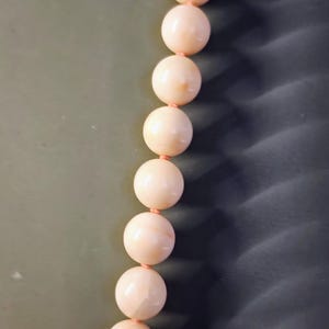 May include: A strand of pale pink coral beads strung together with a thin pink cord. The beads are round and smooth, and they have a slightly iridescent sheen.